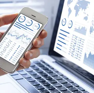 A person is holding a mobile phone with graphs and charts on the screen, and it is placed on top of a laptop with a keyboard and a monitor displaying a world map and financial data.