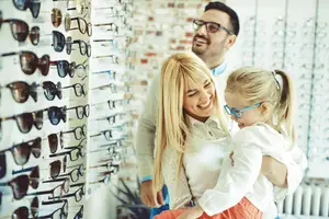 A family is looking at sunglasses in a store, the father is wearing glasses and smiling, the mother is carrying the child, and they are all happy.