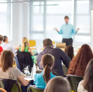 Inside a conference room, several people are seated and listening to a man giving a presentation while gesturing with his hands.