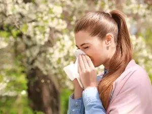 A woman with long brown hair is holding a tissue and blowing her nose in an outdoor area with blurry trees in the background.