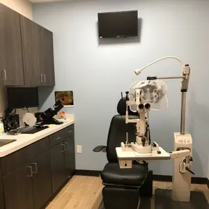 An eye exam chair with an eye exam machine is in a room with blue walls and wooden floors. A monitor is mounted on the wall, and a computer is on the countertop. Cabinets are mounted on the wall, and a sink is on the left. A power outlet is on the wall. A small table with a monitor is in the corner.