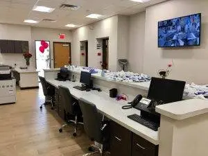 The interior of an office has a white countertop with monitors, keyboards, mice, and telephones. A television is mounted on the wall. There are also chairs and desks. A flower vase is placed on the counter. A printer is on the left side, and a door is at the back. A few doors are in the back. A few balloons are hanging on the wall.