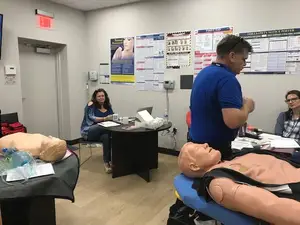 In a room, a man and a woman are standing next to a mannequin on a chair, while the woman is sitting on another chair and looking at a book. A desk is in front of them with a laptop, a bottle, and other items on top. There is a poster attached to the wall, and an exit sign is mounted above the door.