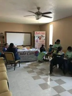 People gathered in a room with a table covered by a banner, some are sitting on chairs and a woman in a wheelchair