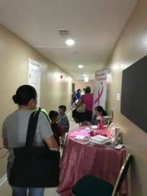 A woman in a gray shirt walks down a hallway with children, passing a table with a pink tablecloth and a pink ribbon banner.