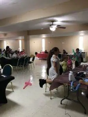 A hall with people seated on chairs around tables, a woman sitting on a chair, a table with items, and a ceiling fan