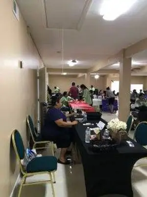 A woman is sitting at a table in a room with many other people, there are other tables and chairs in the room.