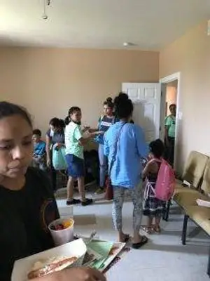 A group of people in a room with a person holding a box of food, others are carrying bags, and a child with a pink backpack