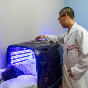 A man in a white lab coat stands next to a woman lying in a blue light bed, both looking at the equipment.