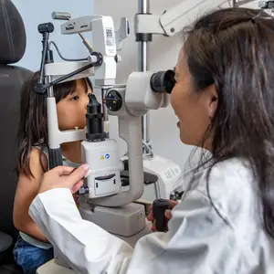 An adult woman examining the eyes of a young girl using an ophthalmoscope in a medical office.