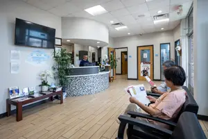 A woman is sitting and reading a magazine while an older man sits next to her and waits for the doctor inside a clinic. The receptionist is working behind the reception desk, with a monitor, potted plants, and a TV mounted on the wall. The clinic has a wooden floor and a glass window with a view of the outside.