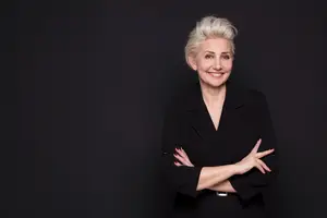A smiling woman with short blonde hair wearing a black blazer poses against a dark background.