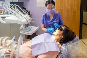 A woman is sitting on a dental chair while a dentist in a blue uniform and face mask is cleaning her teeth.