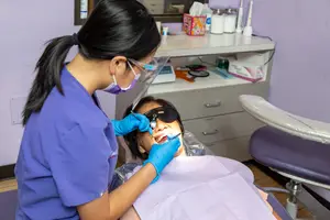 A dentist in blue scrubs and a mask is examining a child's teeth with a dental mirror and probe.