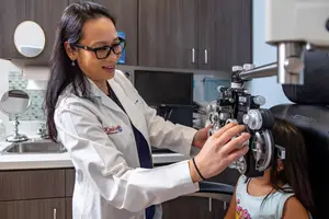 A woman doctor in a white coat examines a young girl's eyes with an ophthalmoscope in an exam room.