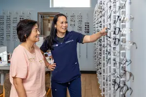 Two women, one in a blue uniform and the other in a pink shirt, are standing in front of a wall with eyeglasses hanging on it, possibly in an optician's shop.