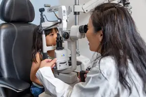 A doctor is examining a young girl's eyes with a slit lamp in a clinic.