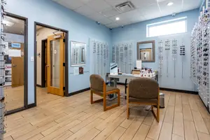 An empty room with blue walls and wooden flooring has a desk with items on it and two chairs in front of a glass door. There are mirrors and frames on the wall and a window with a view of the outside. There are also eyeglasses on the wall and a trash bin on the right side.