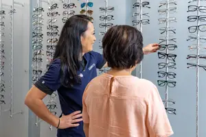 A woman in a blue uniform stands in front of a wall of eyeglasses, showing a pair to a customer.