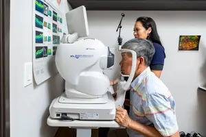 An elderly man is undergoing an eye examination in a clinic while a woman stands beside him, both of them facing the camera.