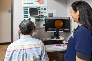 An elderly man is having an eye exam with a female ophthalmologist while looking at a computer screen with an eye image.