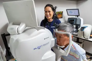 An older man is getting his eyes checked by a woman in an eye clinic with monitors and medical equipment in the background.