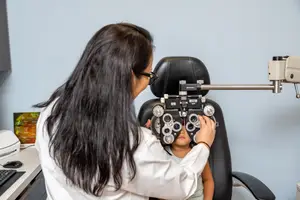 A young girl sits in a chair while a woman, presumably a doctor, uses an ophthalmoscope to examine her eyes.