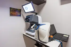A room with a white desk, on which a laptop and an Oculus eye scanner are placed. The eye scanner is connected to a monitor and has a wire attached to it. A poster is on the wall behind the desk.