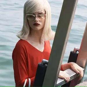 A woman with blonde hair and red lipstick stands by the water, wearing glasses and holding a metal handrail.