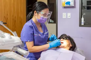 A dental hygienist wearing PPE is cleaning a patient's teeth.