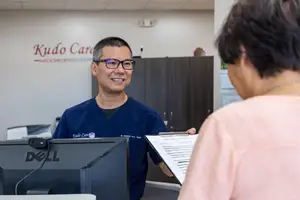 An Asian man wearing a blue shirt and glasses is smiling and holding a clipboard and paper while talking to an older woman in a pink shirt in an office with a white wall and a cabinet with a plant on top and a monitor on the left