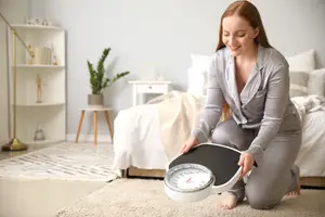 A woman wearing pajamas is kneeling on the floor and holding a digital scale in her hands