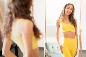 A woman is looking at herself in a mirror, wearing yellow clothing, and smiling.