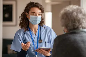 A nurse in a blue uniform and mask speaks with an elderly patient while holding a tablet.