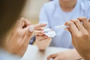 A person is trying to put on a contact lens in their eye while holding a contact lens container in their other hand.