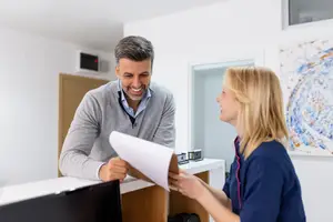 A man and a woman standing in front of a desk in an office room with a white wall, smiling and looking at a piece of paper in the man's hand