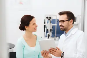 A man and a woman are in a room with an ophthalmoscope behind them. The man is holding a tablet, and the woman is smiling and looking at him. They are both wearing glasses.