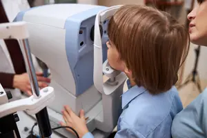A young girl is using an eye exam machine while an adult woman is watching her in a hospital room.
