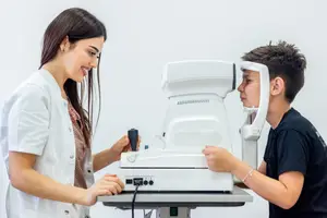 A woman and a boy are standing in front of a white machine with a black wire, and the woman is holding the boy's chin with her left hand while the boy is looking at the machine.