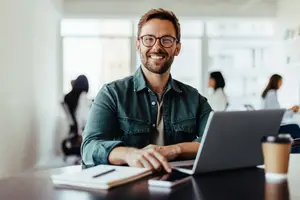 A smiling businessman sitting at a desk with a laptop, a cup, a book, and a pen in an office.