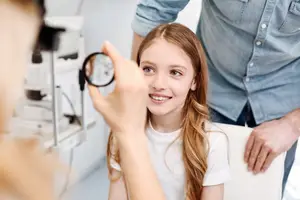 A young girl is getting her eyes checked by a woman wearing a blue shirt and a man wearing a denim shirt in an optical shop.
