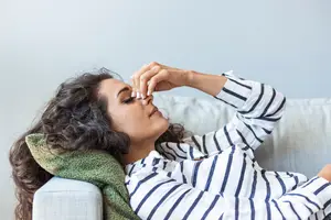 A woman lying on a couch and touching her nose with her right hand, probably blowing her nose, wearing a striped long-sleeve shirt and a green blanket around her neck.
