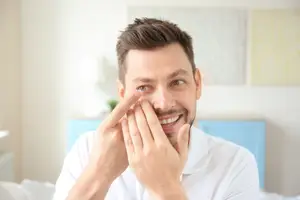 A man wearing a white shirt is putting in a contact lens in his right eye while smiling and looking at the camera.
