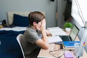 A boy sitting at a desk with his hand covering his face and looking stressed