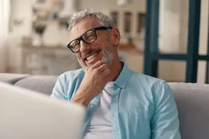 An older man wearing glasses is smiling while sitting on a couch and looking at a laptop.