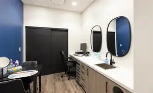 A room with a table, chairs, a monitor, a sink, and mirrors on the wall.