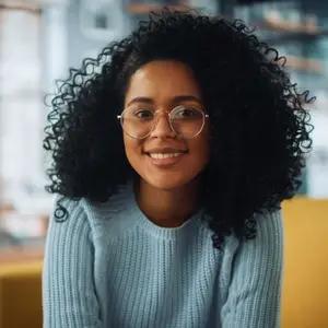 A woman with curly hair wearing glasses and a blue sweater sitting on a couch smiling