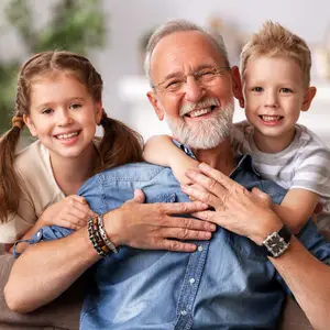 A smiling elderly man sitting on a couch with two children, a girl and a boy, hugging him from behind, with the girl wearing a white dress and the boy wearing a gray shirt, while the elderly man is wearing a blue shirt, a watch, and glasses.