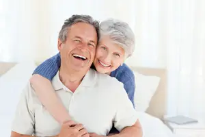 An elderly couple smiling and embracing in a bedroom with white curtains and a bed