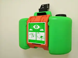 An eye wash station with a green and red body, mounted on a wall, displaying an icon with a green circle and an eye symbol, and two illustrations showing a person washing their eyes and another person using a bottle